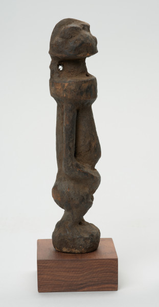 rzeźba - Ujęcie lewej strony rzeźby. Rzeźba w drewnie w formie uproszczonej. Przedstawia postać męską o lekko zarysowanej głowie, szczupłym, długim tułowiu, krótkie nogi ugięte, ręce oparte na kolanach.
