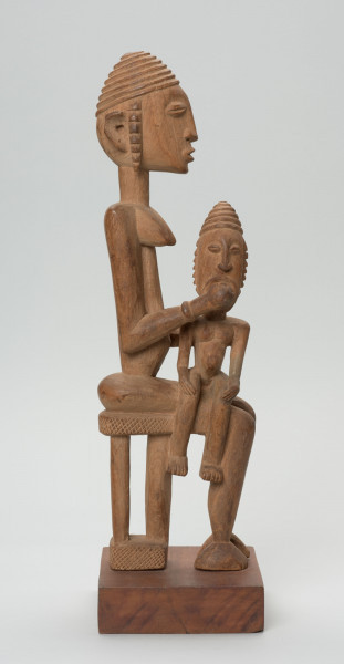rzeźba - Ujęcie z boku; Figurka przedstawiająca postać kobiety siedzącej na stołku i trzymającej na kolanach dziecko. Kobieta ma owalną, wydłużoną twarz oraz długie, zwisające uszy. Rysy twarzy proporcjonalne, z cienkim kształtnym nosem, brwiami oraz wypukłymi oczami i ustami. Dziecko siedzi na kolanach bokiem do kobiety. Kobieta trzyma rękę przy buzi dziecka.