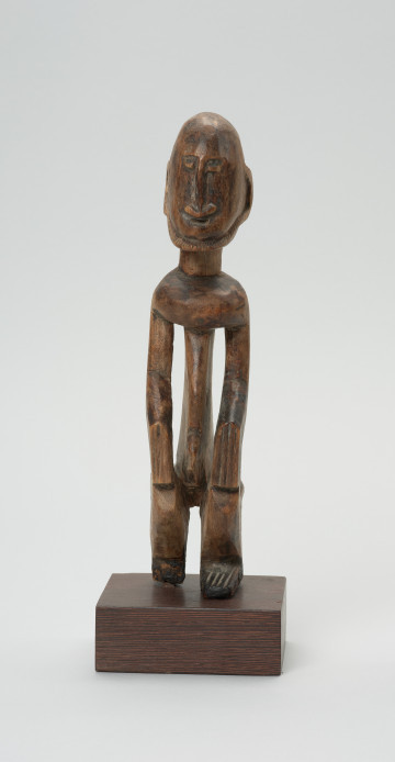 rzeźba - Ujęcie z przodu. Drewniana rzeźba postaci mężczyzny w pozycji siedzącej. Głowa duża z zaznaczonym długim cienkim nosem, strzałkowatym, długie ręce oparte na kolanach, krótkie nogi.