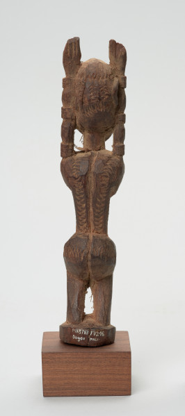 rzeźba - Ujęcie z tyłu. Figura mężczyzny w pozycji stojącej (na ugiętych w kolanach nogach) z rękoma uniesionymi do góry. Cała powierzchnia rzeźby pokryta jest delikatnym rytem, sugerującym skaryfikację.