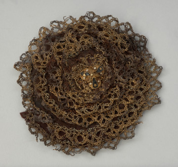 rozeta do obuwia księcia Franciszka I (1577-1620) - Ujęcie z góry pierwszej rozety. Rozeta wykonana z wykończonej złotą koronką jedwabnej wstążki, ułożonej spiralnie i przyszytej do okrągłej podkładki. Spięta pośrodku trójlistnym klejnotem zdobionym barwną emalią. Na odwrocie rozety zachowane fragmenty wstążek.