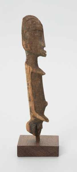 Drewniana figurka kobiety z otworem w lewej piersi - Ujęcie z lewej strony rzeżby. Drewniana figurka postaci kobiecej, na której widoczne są piersi.  Ręce lekko zaznaczone, ułożone wzdłuż tułowia. Brak nóg. Na wysokości lewej piersi otwór.