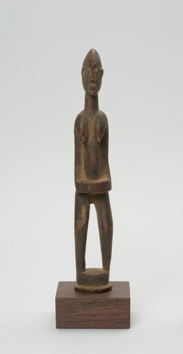 rzeźba - Ujęcie z przodu; Figura - postać kobiety w pozycji stojącej. Głowa mała, podłużna, ręce, piersi, brzuch lekko wydobyte z bryły figury. Nogi bez stóp, przechodzą od razu w okrągły cokolik.