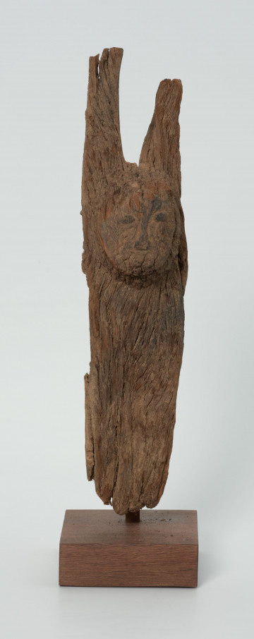 Drewniana figurka na rozwidlonej u góry desce - Ujęcie z przodu; na płaskiej, rozwidlonej na szczycie desce wyrzeźbiono okrągłą głowę z delikatnie zaznaczonymi oczami, nosem z małymi ustami. Figura posiada liczne spękania.