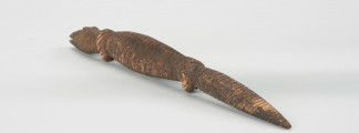 Drewniana figurka krokodyla - Ujęcie z tyłu; Drewniana figurka krokodyla, na całej długości pokryta drobnymi nacięciami. Zaznaczona długa głowa, długi tułów oraz gruby ogon, zakończony spiczaście i spuszczony do dołu. Zaznaczone proste, krótkie kończyny przednie i tylne.