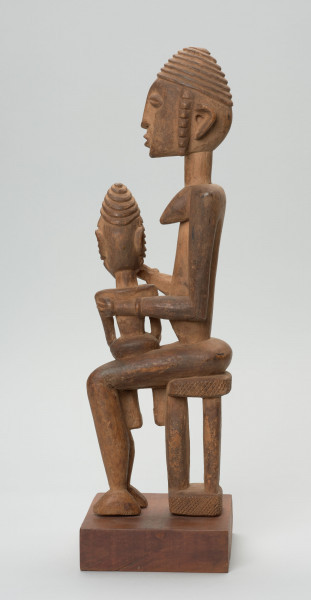 rzeźba - Ujęcie z lewego boku; Figurka przedstawiająca postać kobiety siedzącej na stołu i trzymającej na kolanach dziecko. Kobieta ma owalną, wydłużoną twarz oraz długie, zwisające uszy. Rysy twarzy proporcjonalne, z cienkim kształtnym nosem, brwiami oraz wypukłymi oczami i ustami.