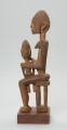 rzeźba - Ujęcie z lewego boku; Figurka przedstawiająca postać kobiety siedzącej na stołu i trzymającej na kolanach dziecko. Kobieta ma owalną, wydłużoną twarz oraz długie, zwisające uszy. Rysy twarzy proporcjonalne, z cienkim kształtnym nosem, brwiami oraz wypukłymi oczami i ustami.