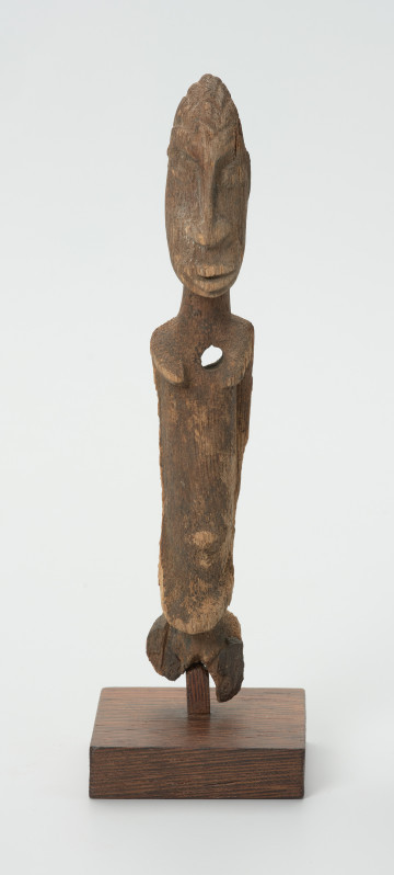 Drewniana figurka kobiety z otworem w lewej piersi - Ujęcie z przodu. Drewniana figurka postaci kobiecej, na której widoczne są piersi.  Ręce lekko zaznaczone, ułożone wzdłuż tułowia. Brak nóg. Na wysokości lewej piersi otwór.