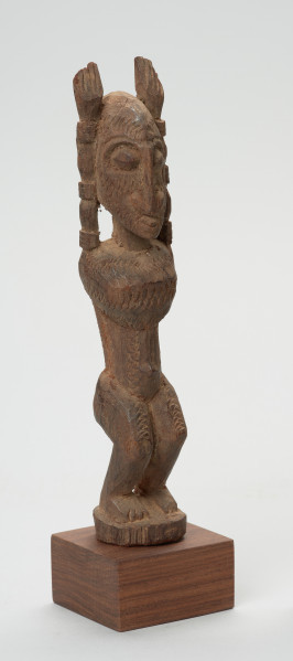 rzeźba - Ujęcie przodu skosem w prawą stronę. Figura mężczyzny w pozycji stojącej (na ugiętych w kolanach nogach) z rękoma uniesionymi do góry. Cała powierzchnia rzeźby pokryta jest delikatnym rytem, sugerującym skaryfikację.