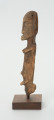 Drewniana figurka kobiety z otworem w lewej piersi - Ujęcie prawej strony rzeźby. Drewniana figurka postaci kobiecej, na której widoczne są piersi.  Ręce lekko zaznaczone, ułożone wzdłuż tułowia. Brak nóg. Na wysokości lewej piersi otwór.