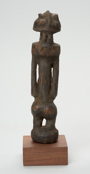 rzeźba - Ujęcie z tyłu. Rzeźba w drewnie w formie uproszczonej. Przedstawia postać męską o lekko zarysowanej głowie, szczupłym, długim tułowiu, krótkie nogi ugięte, ręce oparte na kolanach.