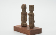 rzeźba - Ujęcie ze skosu z tyłu; Zestaw składa się z dwóch małych, drewnianych figurek przedstawiających postacie ludzkie w pozycji stojącej. Rzeźby o uproszczonej formie i masywnej budowie ciała.