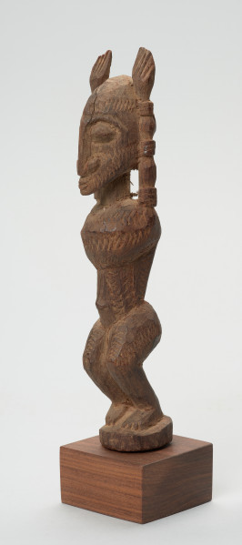 rzeźba - Ujęcie przodu skosem w lewą stronę. Figura mężczyzny w pozycji stojącej (na ugiętych w kolanach nogach) z rękoma uniesionymi do góry. Cała powierzchnia rzeźby pokryta jest delikatnym rytem, sugerującym skaryfikację.