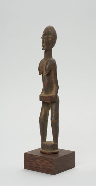 rzeźba - Ujęcie ze skosu z prawej; Figura - postać kobiety w pozycji stojącej. Głowa mała, podłużna, ręce, piersi, brzuch lekko wydobyte z bryły figury. Nogi bez stóp, przechodzą od razu w okrągły cokolik.