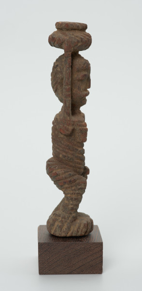 drewniana figura kobiety - Ujęcie lewego boku. Drewniana, rzeźbiona figura kobiety niosącej na głowie naczynie. Postać schematyczna, z uniesionymi ramionami. Głowa jajowata, rysy twarzy uproszczone. Stopy złączone w okrągły cokolik.