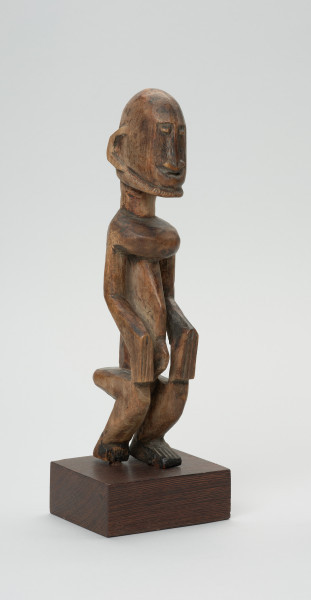rzeźba - Ujęcie przodu skosem w prawą stronę.  Drewniana rzeźba postaci mężczyzny w pozycji siedzącej. Głowa duża z zaznaczonym długim cienkim nosem, strzałkowatym, długie ręce oparte na kolanach, krótkie nogi.