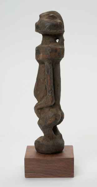 rzeźba - Ujęcie prawej strony rzeźby. Rzeźba w drewnie w formie uproszczonej. Przedstawia postać męską o lekko zarysowanej głowie, szczupłym, długim tułowiu, krótkie nogi ugięte, ręce oparte na kolanach.