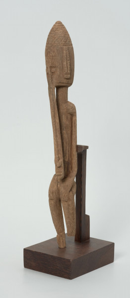 drewniana, rzeźbiona figura - Ujęcie z przodu, z lewej strony. Drewniana, rzeźbiona figura smukłej postaci znajdującej się w pozycji siedzącej. Długa broda ciągnąca się aż do kolan. Prawa dłoń spoczywa na kolanie, lewa trzyma brodę.