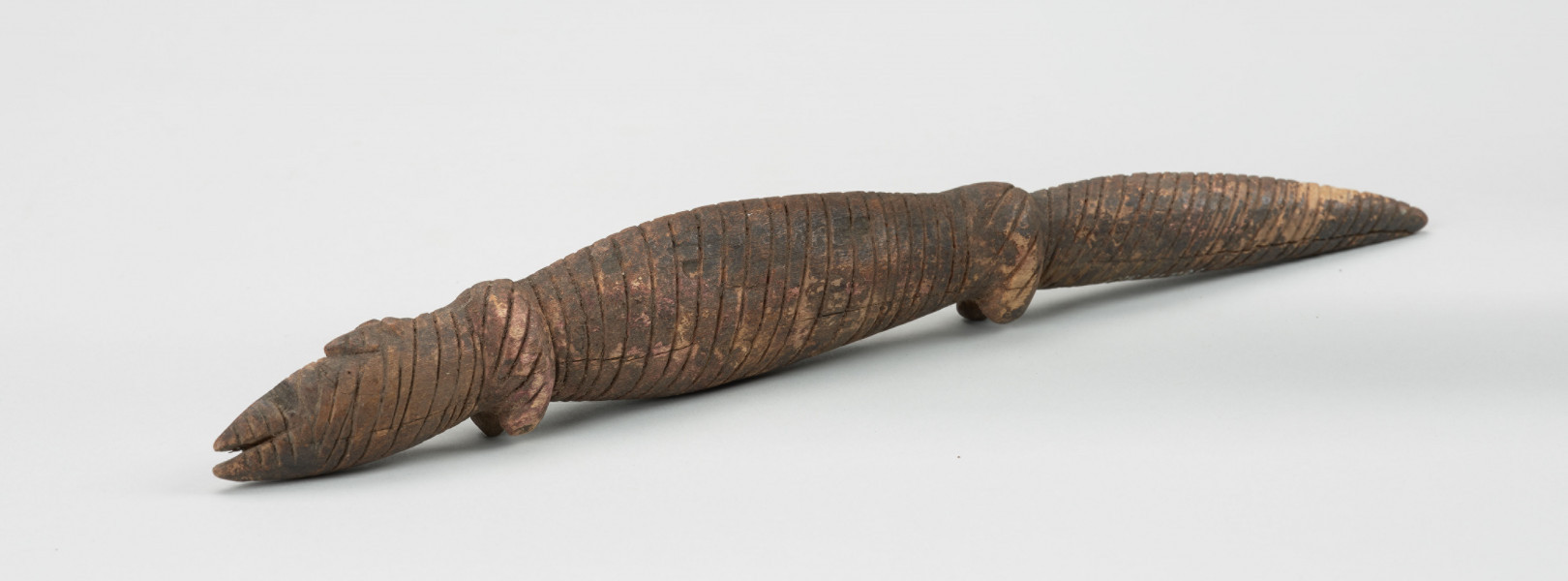 Drewniana figurka krokodyla - Ujęcie z lewej strony z ukosa; Drewniana figurka krokodyla, na całej długości pokryta drobnymi nacięciami. Zaznaczona długa głowa, długi tułów oraz gruby ogon, zakończony spiczaście i spuszczony do dołu. Zaznaczone proste, krótkie kończyny przednie i tylne.