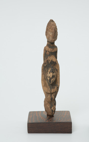 rzeźba - Ujęcie z przodu. Drewniana rzeźba dwóch postaci. Schematycznie zarysowana postać z podłużną głową, szczupłym tułowiem i krótkimi nogami. Większa postać niesie drugą mniejszą z przodu.