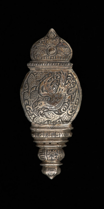 zapinka pendentu, księcia Kazimierza VI/IX (1557-1605) - Ujęcie z przodu. Zapinka służąca do łączenia pendentu z pasem. Dolna część o migdałowym kształcie, zakończonym łukiem w ośli grzbiet, w górnej partii dwa szerokie półwałki oraz jeden mały, wąski, nad którym ozdobnie ukształtowany haczyk. Zapinka w całości pokryta jest grawerowaną, niellowaną dekoracją z motywem ptaka wśród wici roślinnej, stylizowanych kwiatów i ornamentów ciągłych. W dolnej części zapinki mały okrągły otwór na nit.