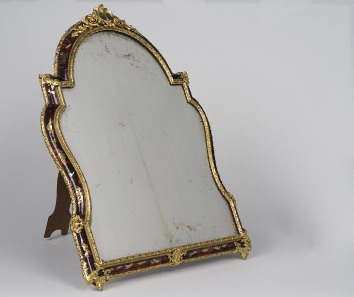 ujęcie 3/4, lustro komody opiera się na podpórce, tafla lustra z przebarwieniami otoczona czarną ramą bogato złoconą.