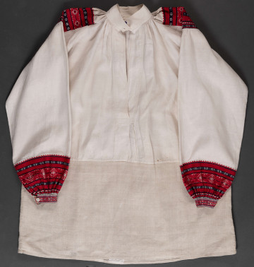 Koszula lniana o kroju przyramkowym, marszczonym. Ozdobiona na przyramkach i mankietach pereborami. Górna cześć koszuli wykonana z płótna lnianego bielonego, dolna – z szarego (dł. 30 cm). Kołnierzyk stójka (4 x 34 cm), zapinany na 3 guziki. Perebory czerwono-granatowe na przyramce (13 x 14,5 cm). Długość pereborów przy mankietach 12 cm. Mankiet (3 x 16 cm) z pętelkami do przeciągania tasiemki, w celu zawiązania.
