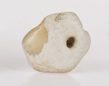 Amulet z przewierconym otworem, kształtu nieregularnego, płaski, od spodniej strony rowek biegnący przez środek. Amulet wykonany ze skały wapiennej (biała kreda)