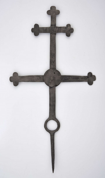 Krzyż żelazny kowalskiej roboty wykonany ze sztabki szer. 2,3 cm i gr. 0,5 cm. Końcówka sztycy, jak i ramion, rozklepana i ukształtowana w formie krzyża o półkolistych ramionach. Górne ramię o szer. 15,7 cm znajduje się w odległości 7,5 cm od górnego zakończenia sztycy. Drugie ramię, dł. 32 cm, przymocowane jest do sztycy nitem, natomiast sposób łączenia zakrywają dwie tarcze o śred. 6,7 cm. W dolnej części sztycy znajduje się otwór o śred. 4 cm. Ostro zakończona sztyca wskazuje na zamocowanie krzyża na przydrożnej figurze lub kapliczce. 