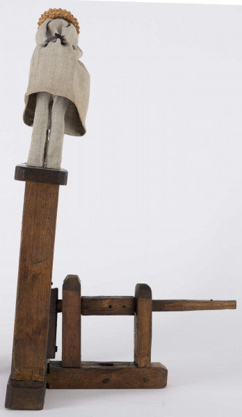 E/2146/ML - Zabawka ruchoma „tracz” wykonana z drewna. Postać odziana w długą płócienną koszulę z rękawami, sięgającą za kolana, plisowana oraz długie lniane spodnie, stóp nie widać. Tracz o schematycznej głowie trzyma długą (46 cm) drewnianą piłę o dużych zębach; piła przechodzi przez podłużny, prostokątny otwór w platformie (wym.: dł. 41 cm, szer. 8 cm, gr. 2 cm), na której stoi tracz. Platforma wsparta jest na dwóch podstawach  (wym.:  wys. 29 cm, szer. 5,5 cm, gr. 2 cm)umocowanych w odległości 10 i 6 cm od krótszych brzegów platformy. Podstawy wpuszczone są w poziomy klocek (wym.: dł. 36 cm, szer. 6 cm, gr. 4 cm), będący podstawą całej konstrukcji. Poniżej 2/3 wysokości dużej piły, umocowana jest mniejsza piła (wym.: dł. 25 cm, szer. 2 cm). Piła ta, prostopadła do dużej, jest ruchoma. Przy niej stał prawdopodobnie drugi tracz. Całość poruszana była korbką (brak) umocowaną do układu drewnianych trybów osadzonych w ramie. Całość połączona na kołeczki, bez użycia gwoździ. 