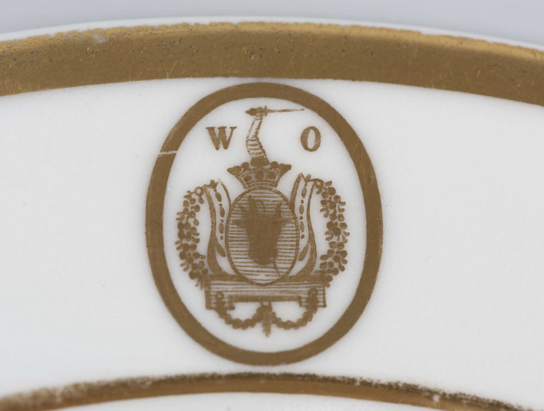 S/CS/1362/1/ML - Talerz płytki, okrągły, gładki, biały, kołnierzujety w dwa złote paski, na kołnierzu złoty owalny medalion z herbem Pomian i monogramem rozdzielonym W O.
Na spodzie sygnatura malowana czerwienią:
 
