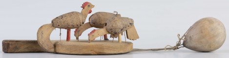 E/17030/ML - Zabawka drewniana przedstawiająca trzy kurki przymocowane do owalnej deseczki z uchwytem. Kurki mają ruchome głowy i ogony, które poruszają się za pomocą sznurków przeciągniętych przez deseczkę i przywiązanych do drewnianej kuli. Deseczka wykonana z drewna sosnowego, kurki z drewna lipowego. Głowy i nogi kurek pomalowane są czerwoną farbą, tułowia zdobi ornament geometryczny w postaci zygzakowatych linii wykonanych brązową farbą.