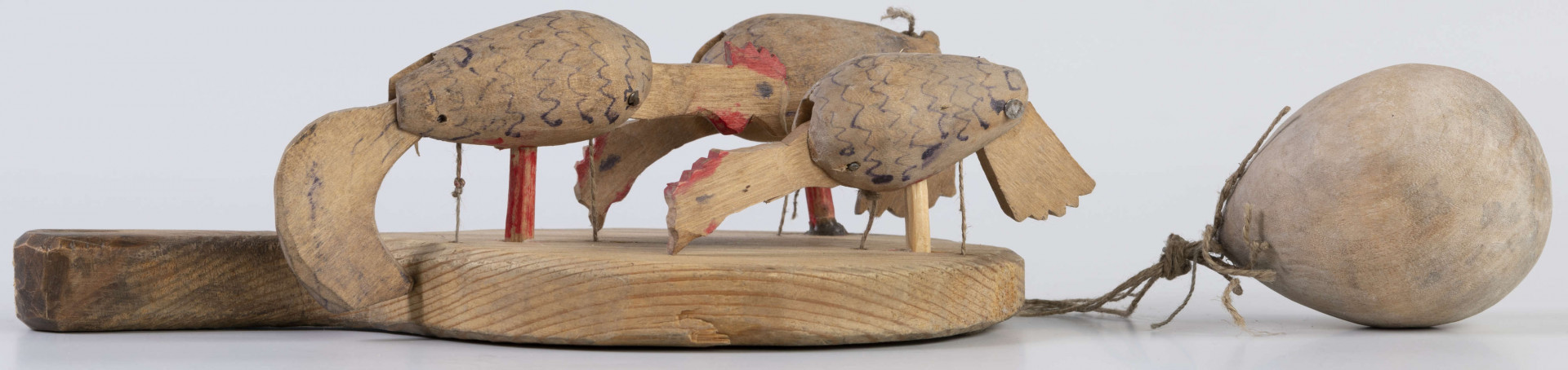 E/17030/ML - Zabawka drewniana przedstawiająca trzy kurki przymocowane do owalnej deseczki z uchwytem. Kurki mają ruchome głowy i ogony, które poruszają się za pomocą sznurków przeciągniętych przez deseczkę i przywiązanych do drewnianej kuli. Deseczka wykonana z drewna sosnowego, kurki z drewna lipowego. Głowy i nogi kurek pomalowane są czerwoną farbą, tułowia zdobi ornament geometryczny w postaci zygzakowatych linii wykonanych brązową farbą.