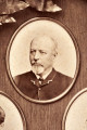 Pośrodku prostokątnego pola o zaokrąglonych narożnikach większa fotografia przyozdobiona wstęgą i gałązkami wawrzynu. Dookoła szesnaście symetrycznie rozmieszczonych mniejszych fotografii. W prawym górnym rogu dr Wacław Lasocki, naczelny lekarz kolei nadwiślańskiej w latach 1875 - 78. W polu ramy w górnej części medalion z płaskorzeźbionym popiersiem z profilu. Przedstawia Leopolda Kronenberga - ojca. Po bokach dwia półleżące aniołki trzymają wieniec laurowy. Po lewej stronie ramy postać robotnika Poleszuka, po prawej pracownika warsztatów kolejowych. W dolnej części front lokomotywy i złoty żeton uprawniający do bezpłatnej jazdy pociągiem I klasy. Fotografia naklejona na tekturę, w dolnej części u dołu sucha pieczęć zakładu fotograficznego.