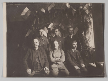 MPol/679/ML - Fotografia przedstawia grupę siedmiu mężczyzn: trzech stojących i czterech siedzących przed nimi. Po środku siedzi Jerzy Pol w mundurze wojskowym. Tło stanowi ściana udekorowana polskimi flagami i zielonymi gałązkami.