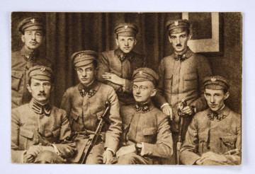 Pocztówka sepiowana - Komenda Naczelna Polskiej Organizacji Wojskowej  w 1914/1915r. (zdjęcie gabinetowe). Ujęcie w poziomie.
AWERS: grupa mężczyzn (7 osób) - podoficerów w mundurach austriackich, z czapkami z umocowanymi orłami legionowymi; trzech żołnierzy trzyma broń białą - szable; postacie w dwóch rzędach - pierwszy siedzący (twarze en face, sylwetki w ujęciu 3/4), drugi stojący; w tle kotara i obraz (?) w ramce .
REWERS: czarna linatura z miejscem na korespondencje; w l. g. rogu nadruk w postaci odznaki pamiątkowej 