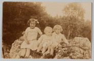 MPol/673/ML - Fotografia przedstawia trójkę dzieci siedzących na skale. Od lewej: Jadwiga Pol (najstarsza), dalej małe dziecko (około 2 lata) i brat Jadwigi, Zbigniew Pol (kilkuletni). Dziewczynka z zaplecionymi po bokach warkoczykami, ubrana w jasną sukienkę, w prawej ręce trzyma drewniany podłużny przedmiot. Zbigniew z długimi, zakrywającymi uszy włosami, ubrany w jasną koszulę i krótkie spodenki, w tle drzewa liściaste.