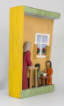 Płaskorzeźba w drewnie lipowym, polichromowana, przedstawiająca wnętrze kuchni, wykonana w jednym kawału drewna. Wnętrze kuchni zaznaczone z lewej strony występem szer. 6,7 cm i gł. 4,5 cm. U góry podkreślony „sufit” wys. 3 cm i gł. 5 cm. Na postumencie wys. 3,5 cm i gł. 4,5 cm. Z lewej strony zakomponowana postać kobiety (z profilu). Kobieta stoi lekko pochylona nad stołem, na którym wałkuje ciasto. Ubrana jest w długą czerwoną suknię w czarne kropki, z długim rękawem.  Na głowie ma zawiązaną, pod brodą, popielatą chustkę, spod której wystają czarne włosy. Twarz płaska, oczy lekko wypukłe, brwi czarne łukowate, nos długi prosty. 
Pod stołem stoi dzbanek, którego brzusiec ozdobiony jest czteropłatkowym kwiatem. Płatki w kolorze czerwony, środek zielony. Wylew podkreślony zielonym paskiem. Z prawej strony przy niskim brązowym stołku, po obu stronach stoją dzieci, które z jednej miski jedzą obiad. Z lewej strony stołka postać chłopca (wys. 12,5 cm) w szarych spodniach, sięgających za kolana i w zielonej koszuli wypuszczonej na spodnie. Twarz chłopca płaska, włosy czarne sięgające do ramion. W prawej ręce chłopiec trzyma łyżkę.  Z prawej – dziewczynka (wys. 13,6 cm), w długiej czerwonej sukience w żółto-czarne ciapki. Włosy czarne, sięgające ramion, twarz płaska. W prawej ręce trzyma łyżkę zanurzoną w misce, lewa opuszczona swobodnie. 
Na wys. 15,5 cm, z prawej strony, okno z kwiatami na parapecie i zasłonkami po bokach, do połowy okna. Wym. okna: wys. 14,5 cm, szer. 11,6 cm.
