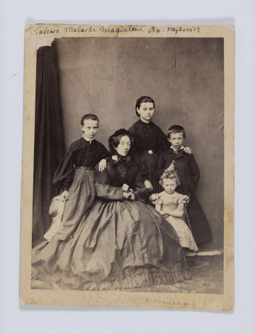 ML/H/F/70/1 - Fotografia przedstawia pięć osób: siedzącą kobietę w wielu dojrzałym, ubraną w ciemną suknię i czepek. Obok niej stoją dwie młode dziewczyny, również w ciemnych sukniach, bez nakryć głowy. Starsza opiera rękę na ramieniu chłopca. Przy starszej kobiecie siedzi mała dziewczynka z jasnymi włosami, ubrana w krótką, jasną sukienkę. Po lewej stoi dziewczyna z włosami zaczesanymi do góry. Jedna ręką opiera się o plecy siedzącej kobiety, w drugiej trzyma słomkowy kapelusz z kokardą. U góry na passepartout napis czarnym atramentem: 