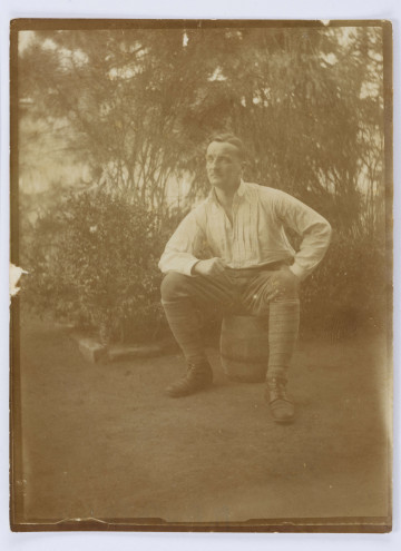 Fotografia przedstawia Jerzego Pola, siedzącego na beczce, z opartymi na kolanach rękoma. Mężczyzna w średnim wieku, ubrany w białą koszulę, ciemne spodnie wpuszczone w zawiązywane trzewiki. W tle widoczne liczne krzewy i drzewa. Fotografia obwiedziona białą ramką.
