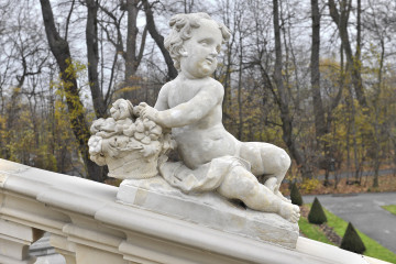 ujęcie od strony pierwszej. Nagie i pulchne dziecko siedzi z w pozycji półleżącej z nogami skierowanymi w prawą stronę.  Z lewej strony przytrzymuje ono kosz z kwiatami. Włosy dziecka są krótki i kręcone.