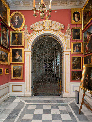 ściana północna, centralnie portal z zamkniętą kratą ozdobiony w górnej części herbem Pilawa. Ściany czerwone (róż pompejański) wiszą na nich obrazy w złotych ramach, podłoga w szare czworokąty.