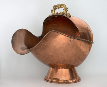 Ujęcie 3/4. Widok na przód i bok. Obiekt umieszczony na stopce o kształcie ściętego stożka. Pojemnik ma kształty zaokrąglone, z szerokim otworem o mocno wygiętej dolnej krawędzi po lewej stronie. Pojemnik i podstawa wykonane z miedzi, o kolorze jasnobrązowym. Na górze, przy krawędzi, widoczna rączka w kolorze złotym.
