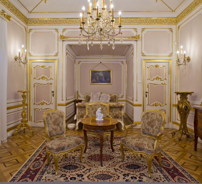 inwentaryzacja wystroju wnętrz, ściana wschodnia, biało-różowe ściany ozdobione złoconymi ornamentami, w centralnej części wnęka z łóżkiem, na pierwszym planie stolik na 3 nogach i dwa krzesła, u góry żyrandol.