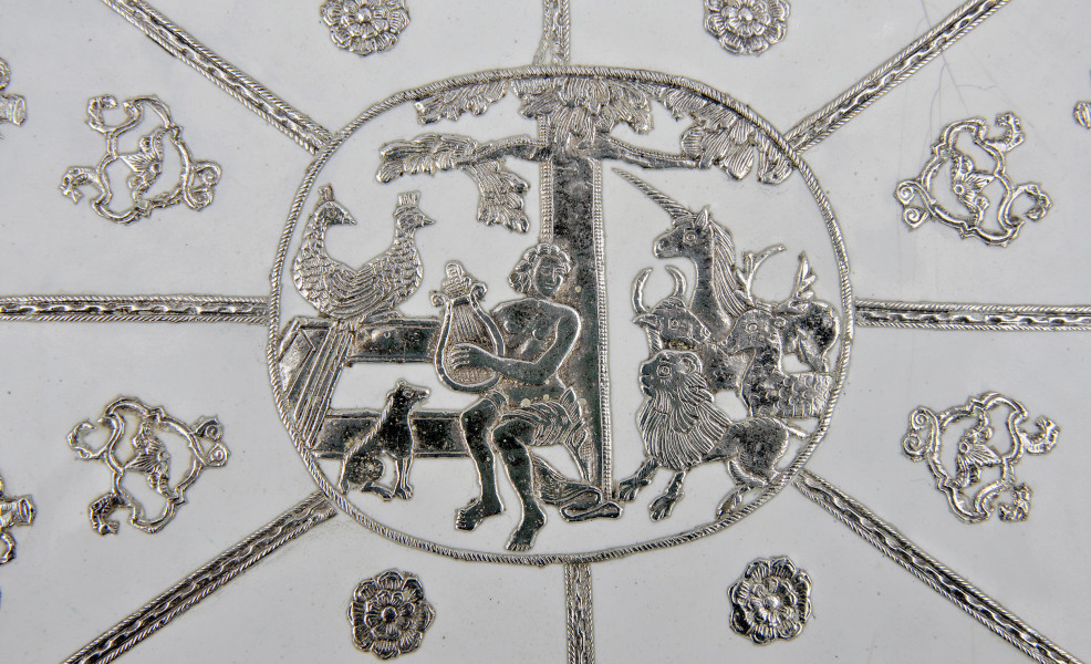 detal awersu - dekoracja srebrna przedstawiająca Orfeusza w otoczeniu zwierząt. Scena w owalu. Na białym tle siedząca postać młodego, półnagiego mężczyzny, opartego o pień drzewa i grającego na lirze. Po obu jego stronach zasłuchane zwierzęta: jednorożec, byk, jeleń, lew, pies i dwa pawie.