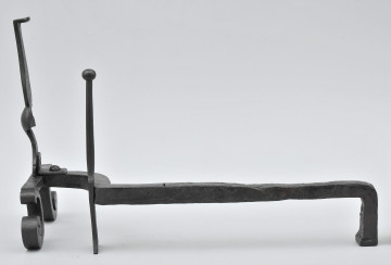 Ujęcie ze wspornikiem na polano. Widok z boku, z przodem po lewej stronie. W większości pręt prosty, bliżej przodu wygięty w prawo. W miejscu wygięcia smukły szpikulec. Pręt łączy się z przednią częścią obiektu przy metalowej płytce. Powyżej niej pionowy trzpień. Poniżej zwężenia na trzpieniu widoczne ubytki po obu stronach. Od płytki odchodzą wygięte stopki. Obiekt w całości czarny, lekko błyszczący.