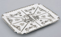 ujęcie ¾. Płaska, prostokątna tacka, pokryta białą emalią i srebrnymi aplikami. W centrum lustra przedstawienie w owalu, reszta podzielona symetrycznie na 8 pól. Brzegi tacki profilowane w narożach i dekorowane srebrnym ornamentem.