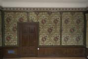 malarsko-sztukatorska dekoracja ściany W 67M0009, ścina obita tkaniną, od dołu cokół na wzór boazerii, po lewej zamknięte drzwi drewniane.
