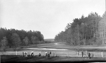 Fotografia czarno-biała przedstawia grupę osób stojących przy drewnianym ogrodzeniu. W tle mokradło i las. 