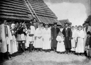 Fotografia czarno-biała przedstawia grupę weselną w strojach ludowych. Mężczyźni, kobiety i dzieci stoją w półokręgu, w tle widoczne strzechy. 
